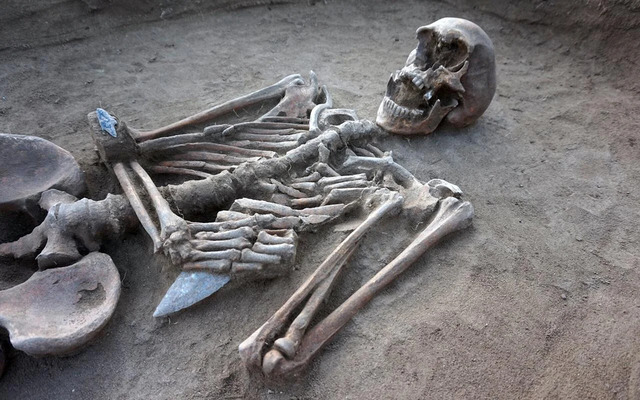 Close-Up Details Show a Skeleton Grasping a Flint Blade in Its Fingers, With an Arrowhead Resting Near Its Elbow.