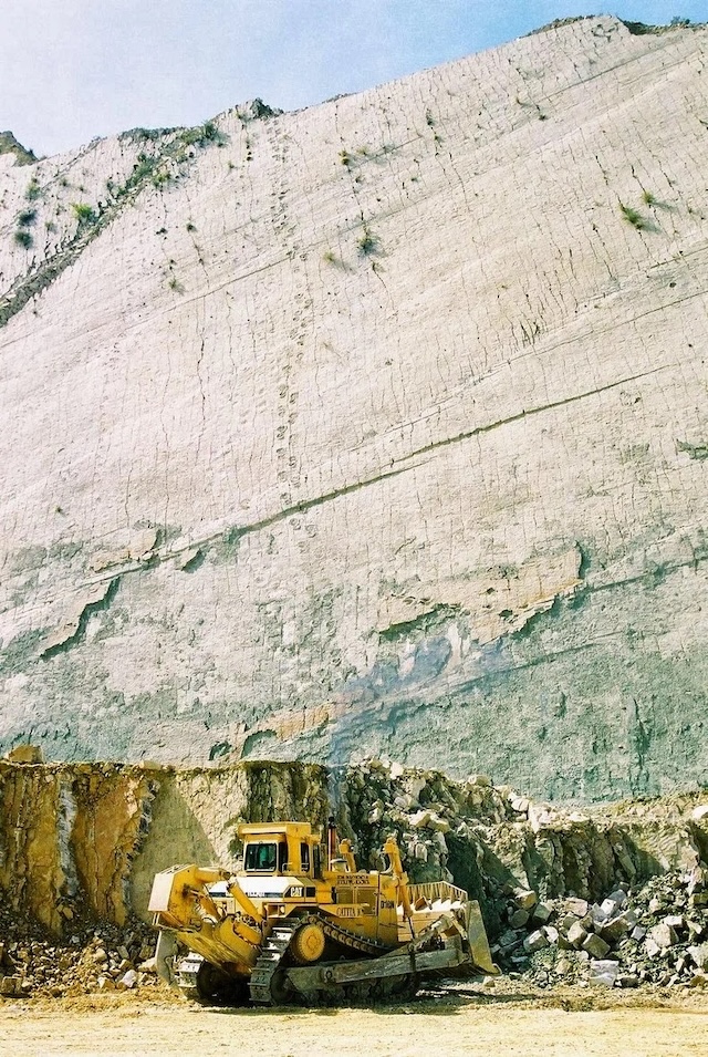 A construction vehicle dwarfed by the immense limestone wall at Cal Orcko, marked with dinosaur footprints left millions of years ago