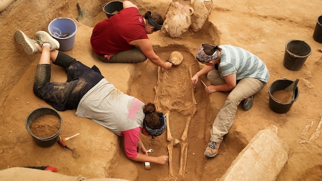 Archaeologists meticulously uncovering a skeleton during an excavation, taking care to preserve both bones and burial artifacts