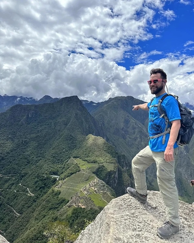 A hiker proudly points out the Machu Picchu site from the top of Huayna Picchu, illustrating the rewarding experience after a challenging climb