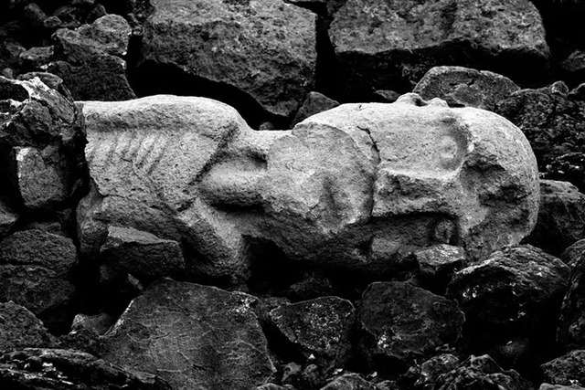 A stone sculpture depicting a humanoid figure found among ancient ruins, showcasing intricate carvings of its face and upper body.