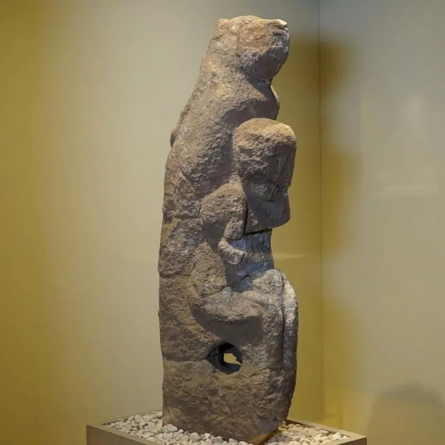 A tall stone statue portraying a human figure with another smaller figure beneath, symbolizing ancient artistic expression.