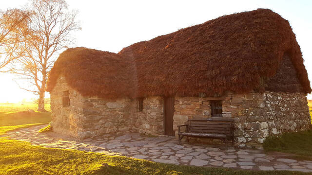 Old Leanach Cottage glowing under the warm hues of a setting sun, with its thatched roof adding to its rustic charm.