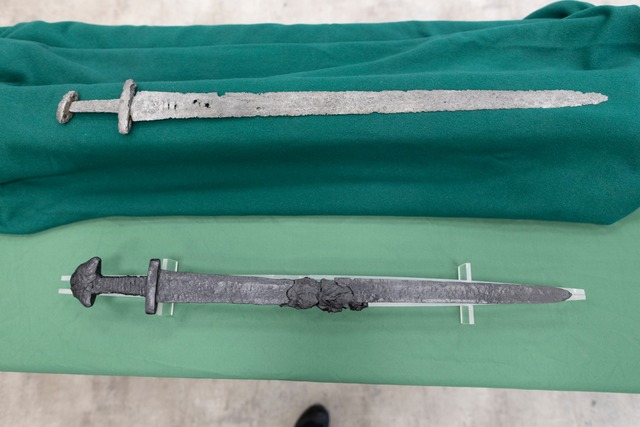 Two Viking-era swords displayed on a green cloth, highlighting the exceptional preservation of the recent discovery alongside another historic artifact.