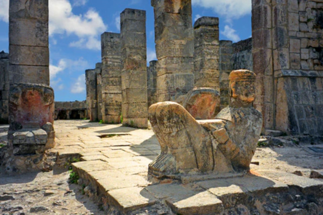 A Chac Mool figure is situated within an ancient ceremonial site, surrounded by stone columns that hint at its integral role in religious practices of the time.