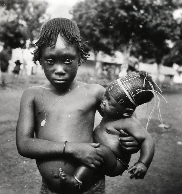 A young Mangbetu girl holding her baby sibling, whose head wrapping reflects the ongoing practice of skull elongation in the family.