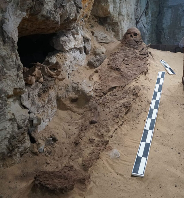 A mummified individual lying in a burial shaft, with skeletal remains of other individuals and artifacts in the background. Credit: Ministry of Tourism and Antiquities of Egypt.