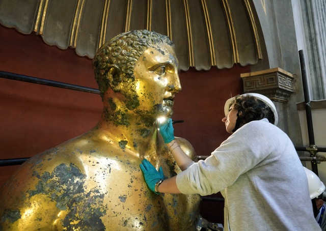 A restorer carefully examines the statue's delicate gilded surface under magnifying tools to preserve its original brilliance.