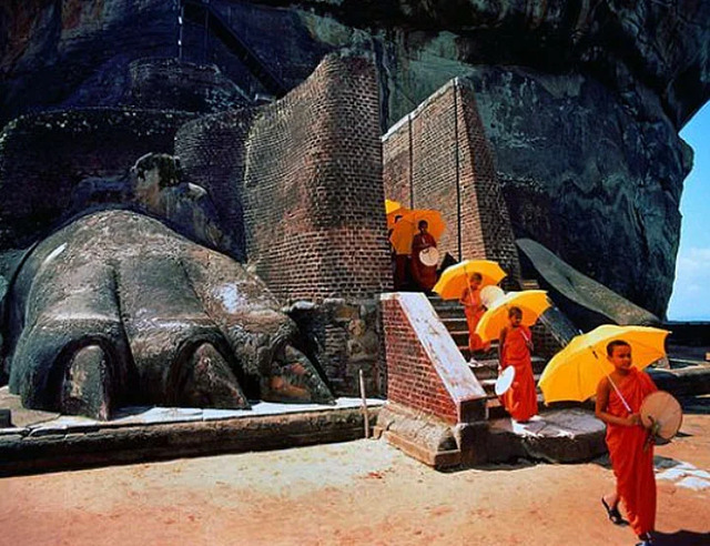 Buddhist monks adorned with vibrant yellow umbrellas walk past the Lion Paws Gate, emphasizing the site’s enduring cultural and spiritual relevance.