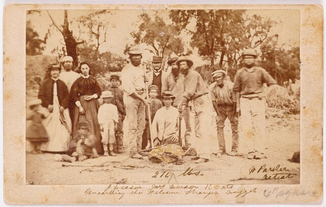 Miners and their wives proudly pose with the finders of the Welcome Stranger nugget, Richard Oates, John Deason, and his wife, capturing a moment of triumph.