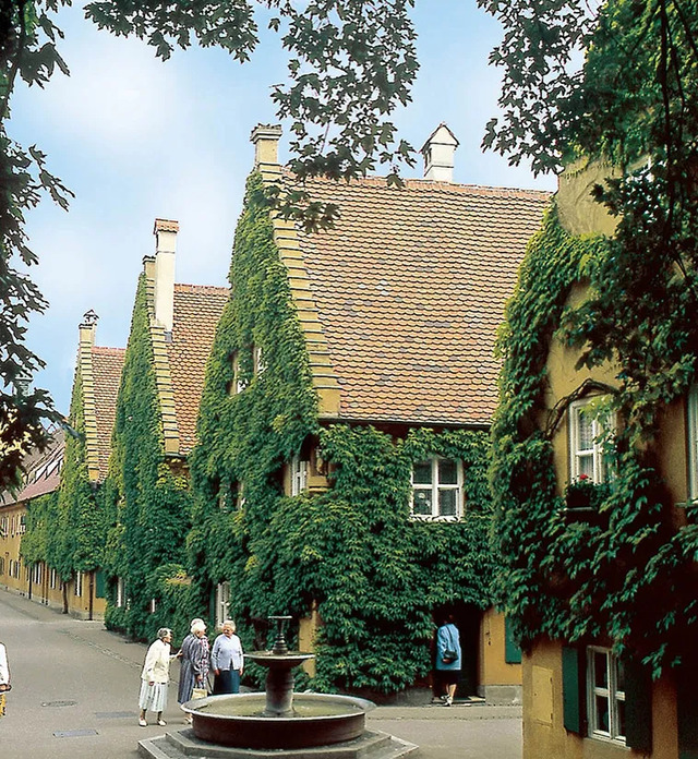 Residents and visitors stroll along the picturesque streets of Fuggerei, framed by charming yellow houses adorned with lush greenery.