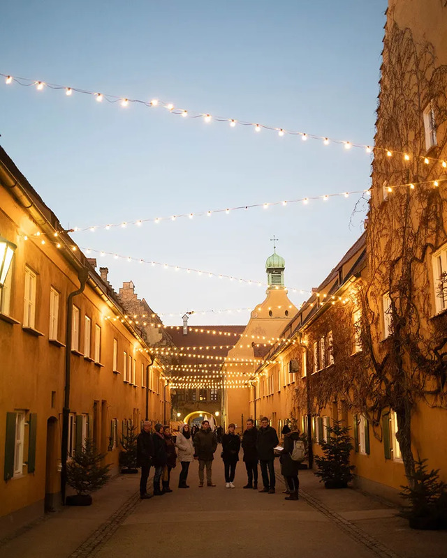 Warm string lights illuminate the historic streets of Fuggerei during an evening celebration, creating a magical atmosphere for visitors and residents alike.